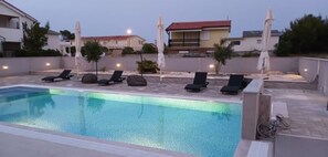 A swimming pool with lounge chairs and umbrellas, surrounded by a low wall and houses in the background. - Apartments Sana (Vir)