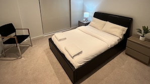 A modern bedroom with a large bed, a chair, a bedside table with a lamp, and a closed sliding door. - Impeccable 1bed Apartment in the Heart Ofgreenford (Greenford)