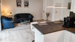 A modern living room with a dark blue sofa, a glass coffee table, and a white armchair. There is a kitchen area with a built-in oven and a coffee machine.
