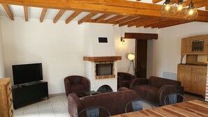 A cozy living room with a fireplace, wooden ceiling beams, a TV, and a dining area with a wooden table. - Gîte La Longère Proche de Saint-valéry, au Coeur de la Baie de Somme (Saigneville)