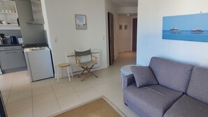 Un soggiorno con un divano grigio, un tavolino, una sedia pieghevole e un angolo cottura con frigorifero e fornello.