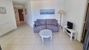 Un soggiorno con un divano grigio, un tavolino da caffè bianco, una TV su un mobile e un quadro raffigurante una costa.