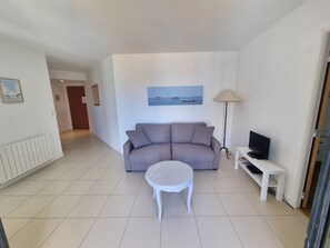 Un salon avec un canapé gris, une table basse blanche, un téléviseur posé sur un meuble et un tableau représentant un paysage côtier.