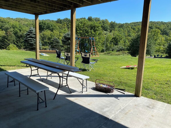 An outdoor patio area with picnic tables and chairs, a swing set, and a grassy field with trees in the background. - 5 minutes to Penn State sporting events. (Port Matilda)