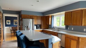 Une cuisine moderne avec des meubles en bois, un îlot central doté de tabourets, un réfrigérateur et une fenêtre laissant entrer la lumière naturelle.