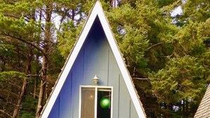 A triangular house with a small table and two windows, surrounded by trees. - Mermaid's Rest  Oceanfront A Frame  Pet Friendly (Otter Rock)