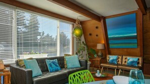A living room with a leather sofa, a dining table with a blue tablecloth, and a painting of a beach scene. - Mermaid's Rest  Oceanfront A Frame  Pet Friendly (Otter Rock)