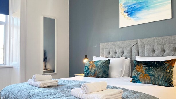 Un dormitorio moderno con cabecera gris, un cuadro abstracto grande y toallas ordenadas sobre la cama.