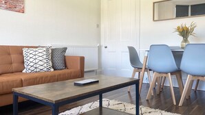 Una sala de estar con un sofá marrón, cojines estampados, una mesa de centro y una zona de comedor con una mesa y sillas.