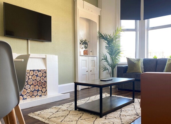 A living room with a flat-screen TV mounted on the wall, a fireplace with a decorative log pattern, a large potted plant, and a sofa with cushions. - ByEvo 19 Walker GLA Apartment (Paisley)
