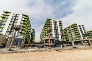 A modern multi-story residential building with balconies, a medical facility, and a landscaped area with benches and palm trees. - Apartamente Indigo Alezzi (Navodari)