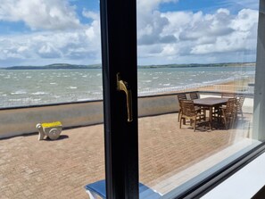 On peut voir, à travers une fenêtre, une terrasse avec une table et des chaises, d’où l’on aperçoit une étendue d’eau et une côte lointaine.
