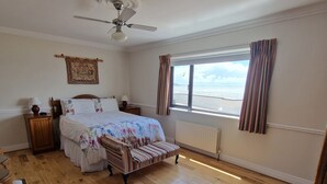 Quarto com cama, cadeira, mesa de cabeceira, janela com cortinas, ventilador de teto e radiador.