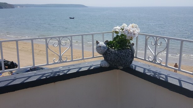 Uma varanda com um vaso decorativo de flores brancas, com vista para uma praia e um mar calmo, com um barco ao longe.