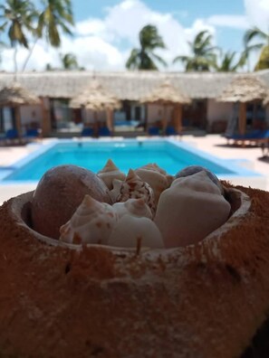Une piscine au bord de la plage, avec des chaises longues et des parasols, des palmiers en arrière-plan et un bol de crème glacée au premier plan.
