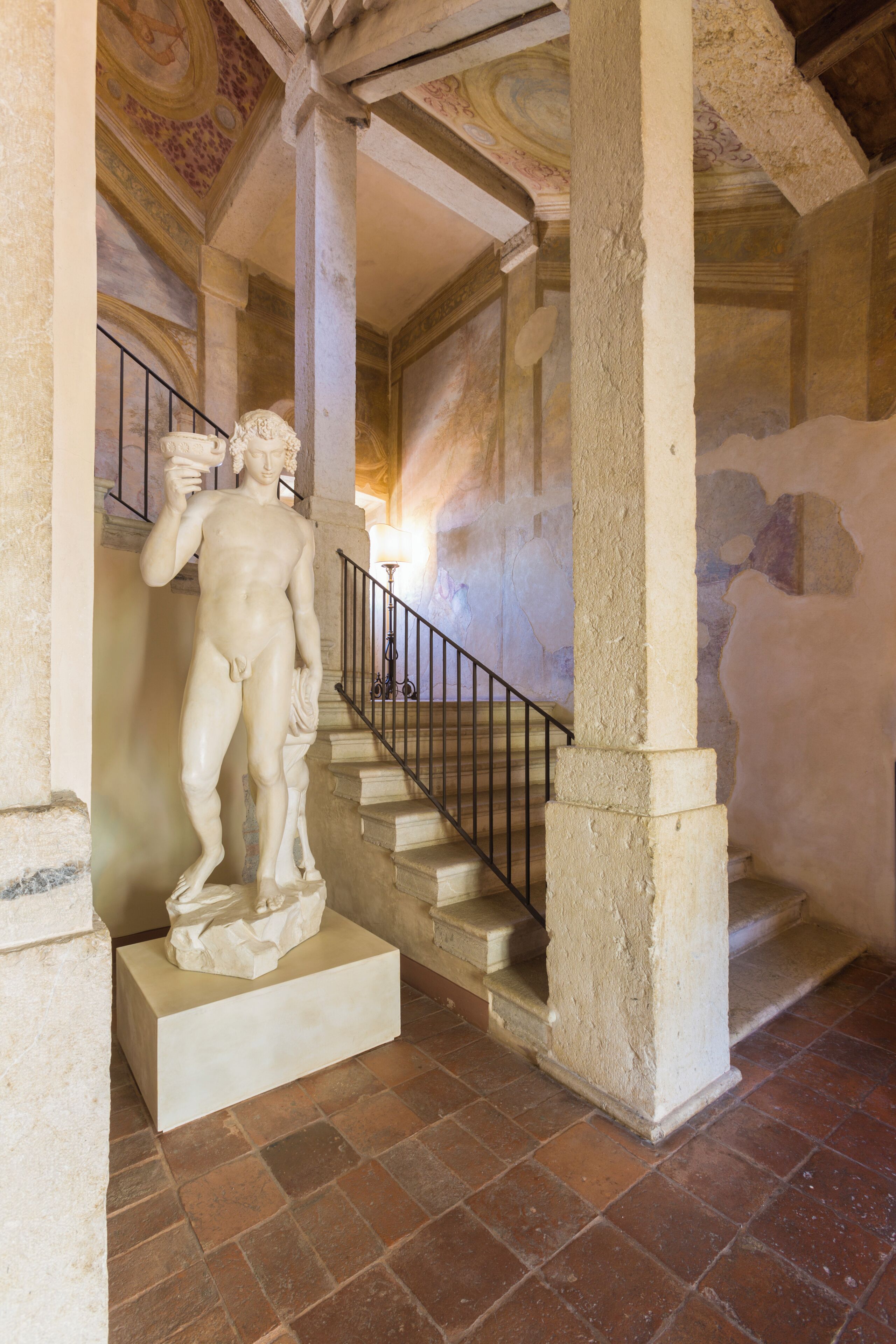Une statue de marbre, posée sur un piédestal, dans un bâtiment doté d’un escalier et d’un sol carrelé.