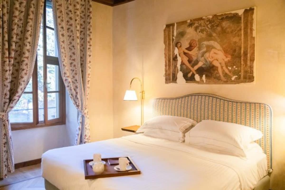 A neatly made bed with a tray of breakfast items, a painting on the wall, and a window with curtains.