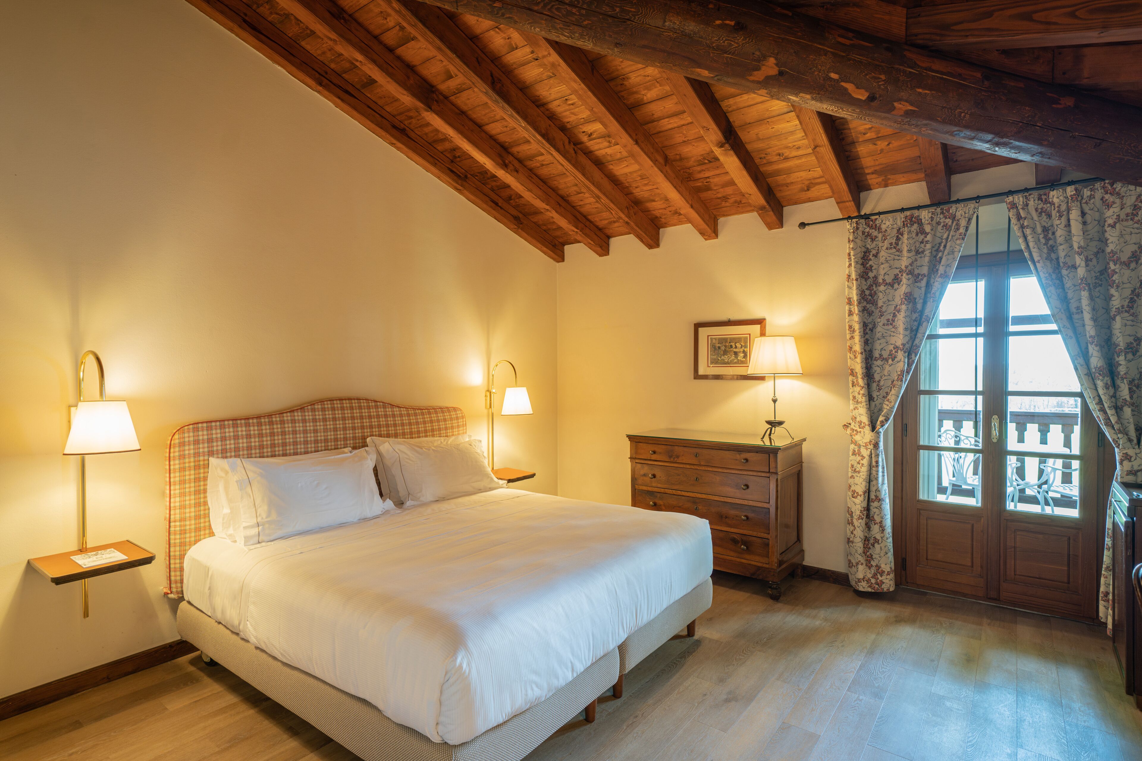 A bedroom with a wooden ceiling, a bed with white bedding, two bedside lamps, a wooden dresser, and a window with curtains.