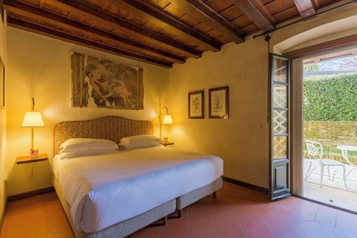 A bedroom with a bed, two bedside lamps, a painting on the wall, and a sliding glass door leading to an outdoor area with a table and chairs.