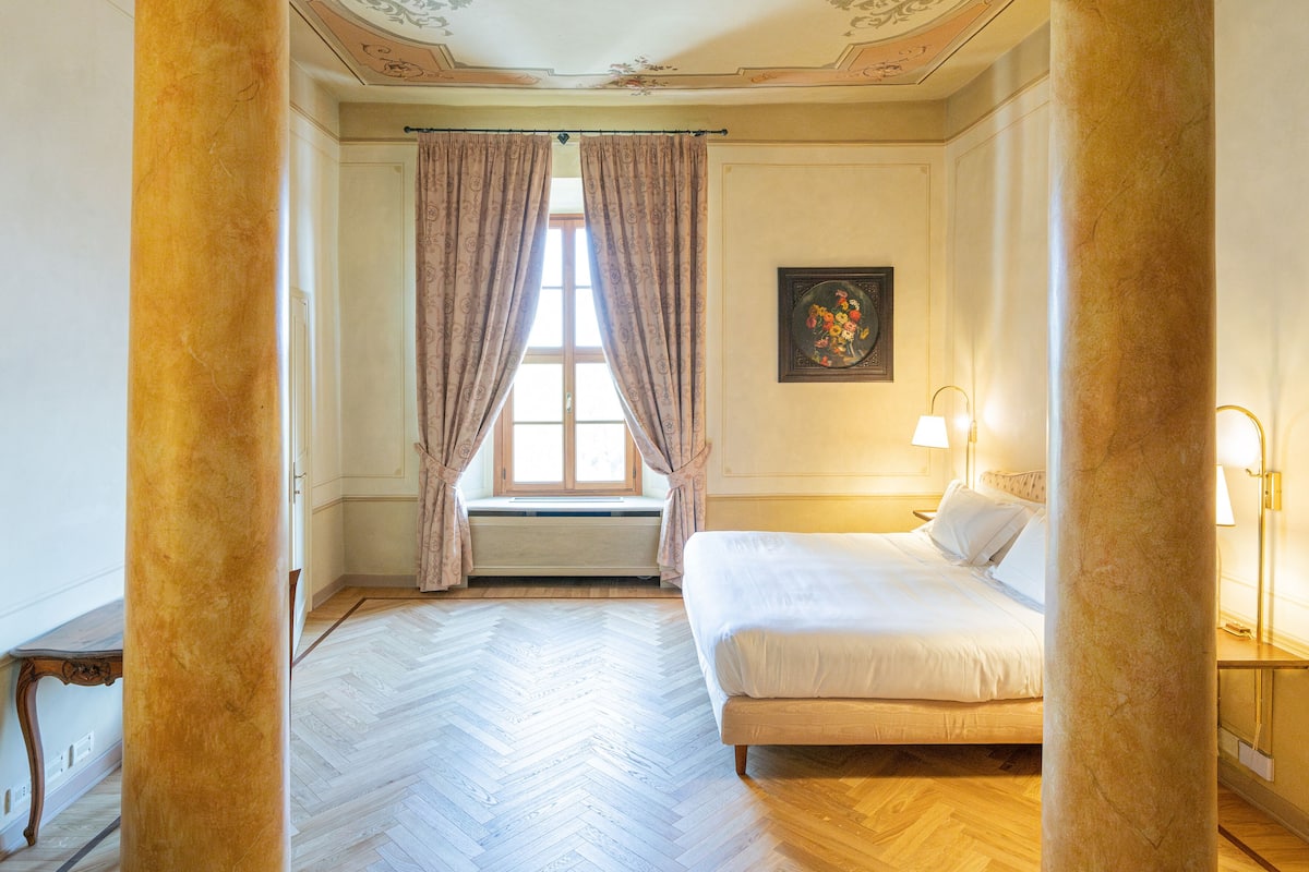 Une chambre d’hôtel avec un grand lit, une fenêtre avec des rideaux, un tableau au mur et un plancher en bois.