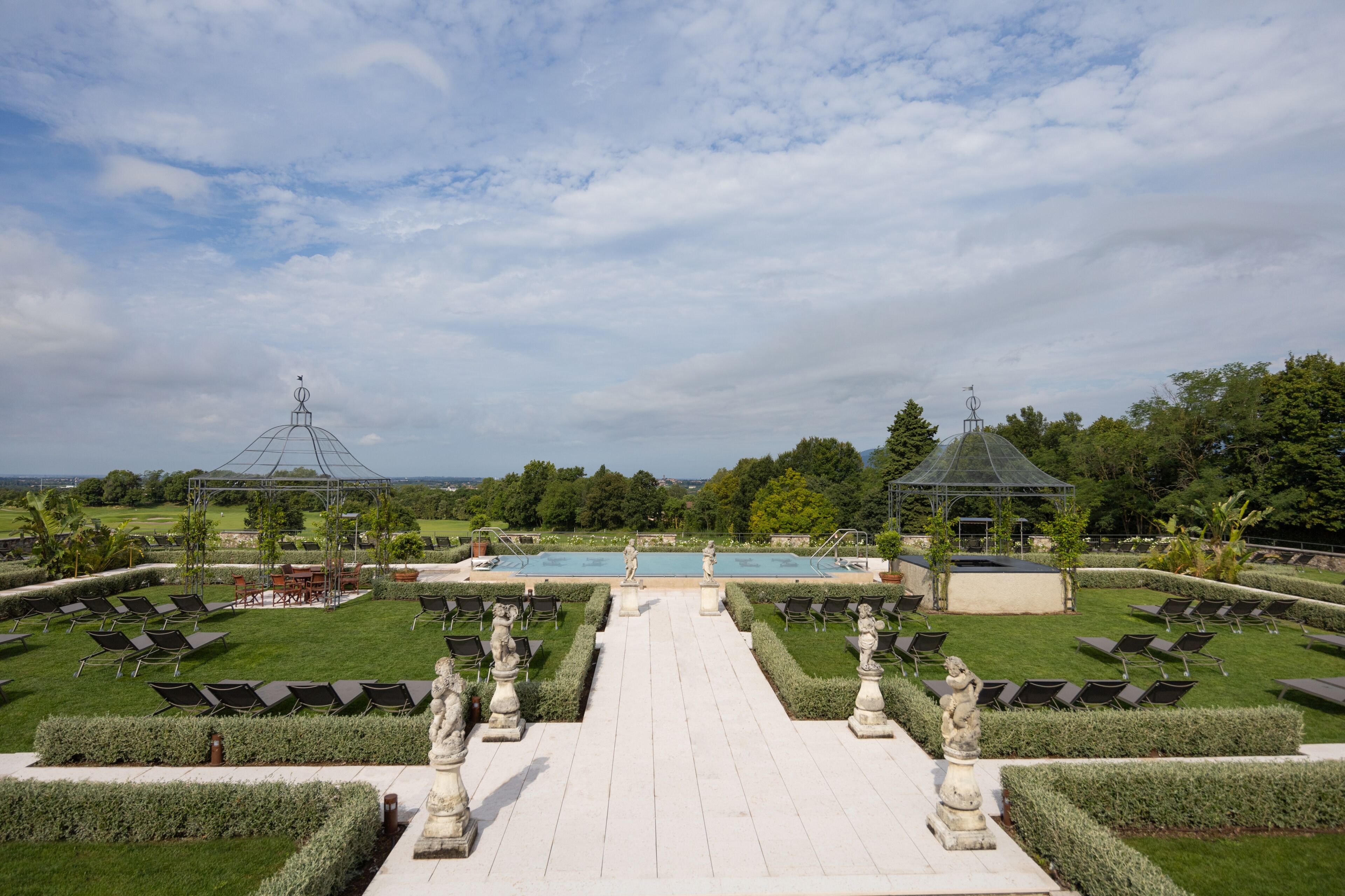 Un jardín formal con un sendero central que lleva a una piscina, rodeado de estatuas y setos cuidados.