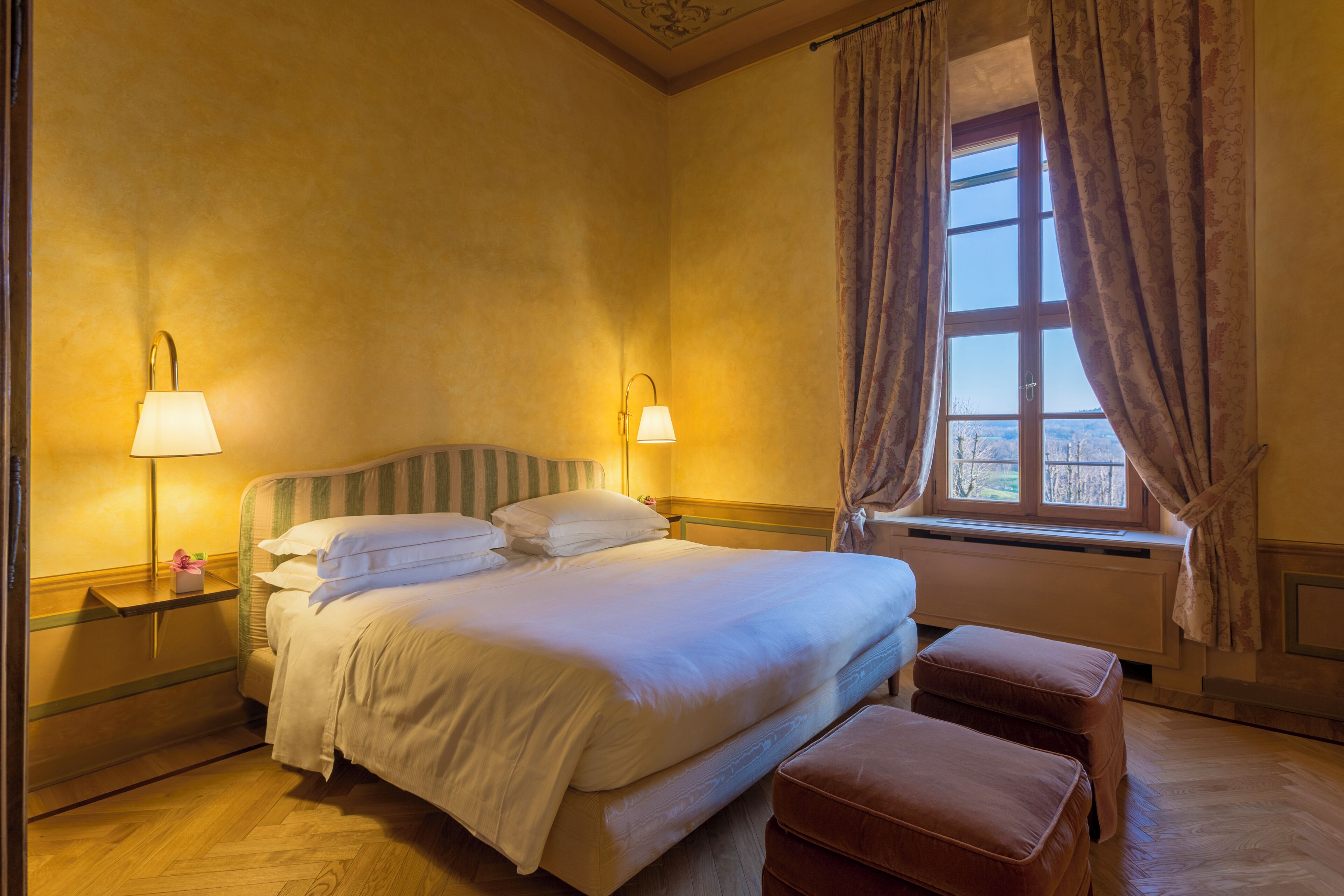 A hotel room with a bed, bedside lamps, a window with curtains, and a view of the outdoors.
