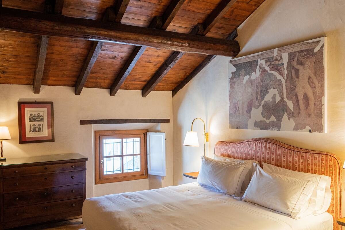Une chambre avec un plafond en bois, un lit avec une literie blanche, une table de chevet, une lampe fixée au mur et une fenêtre avec un cadre en bois.