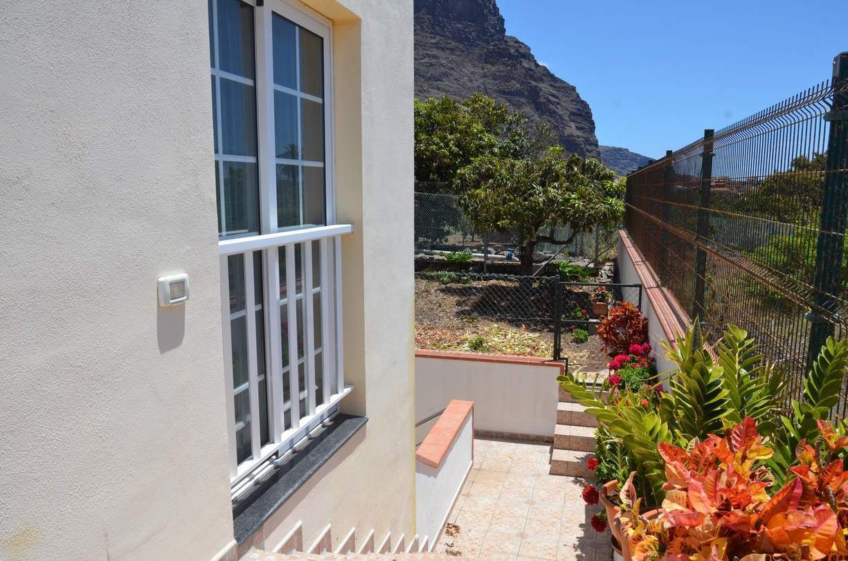 A balcony with a view of a mountainous landscape, a fenced garden area, and a building with a white exterior and a window with white frames.