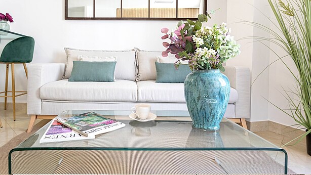 Ett modernt vardagsrum med ett soffbord i glas, en vit soffa med turkosa kuddar, en blå vas med blommor och en tidning.