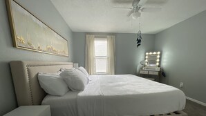 Una cama bien tendida con sábanas blancas, una mesita de noche con lámpara, un ventilador de techo y un espejo grande con luces.