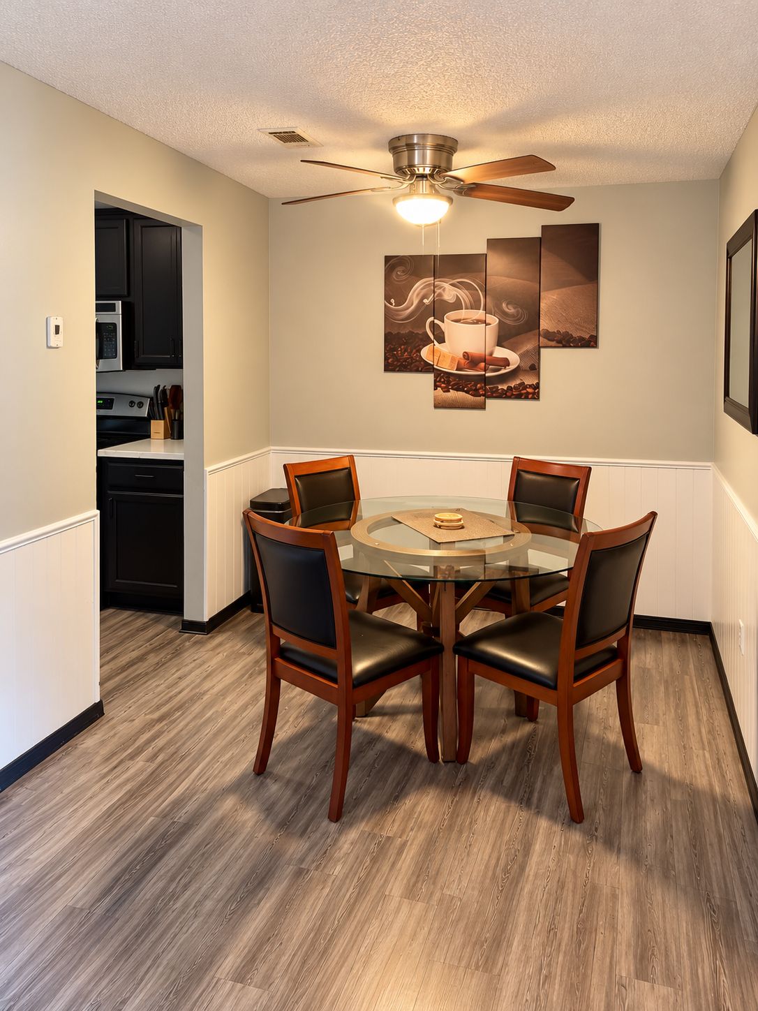 Un coin repas avec une table ronde en verre, quatre chaises en bois, un ventilateur de plafond et, au fond, une cuisine.