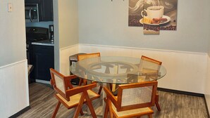 Un comedor con mesa de vidrio rodeada de sillas de madera con asientos de mimbre, un ventilador de techo y una obra de arte en la pared que representa una taza de café.