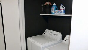 Un cuarto de lavandería con lavadora y secadora de carga frontal, un estante con productos de limpieza y una puerta blanca.