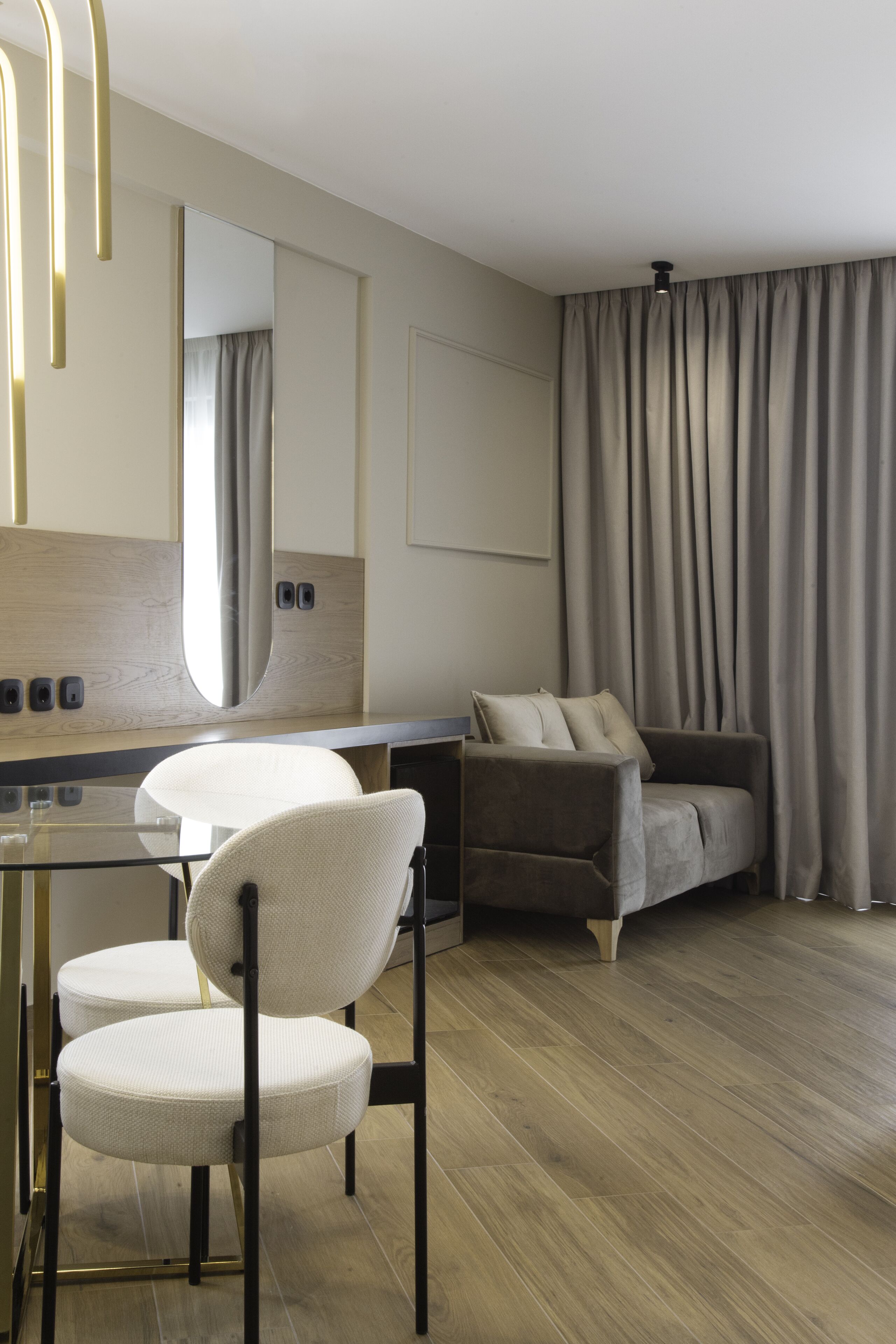 Une chambre d’hôtel moderne avec un plancher en bois, un bureau en verre, une chaise blanche, un canapé gris et une grande fenêtre avec des rideaux.