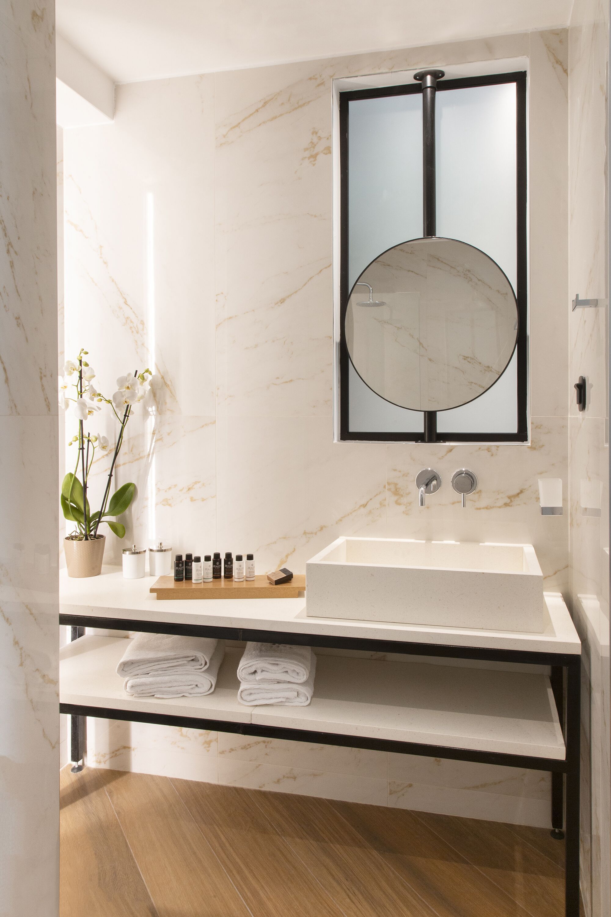 A modern bathroom with a marble sink, a large mirror, a potted orchid, and neatly folded towels on a shelf.