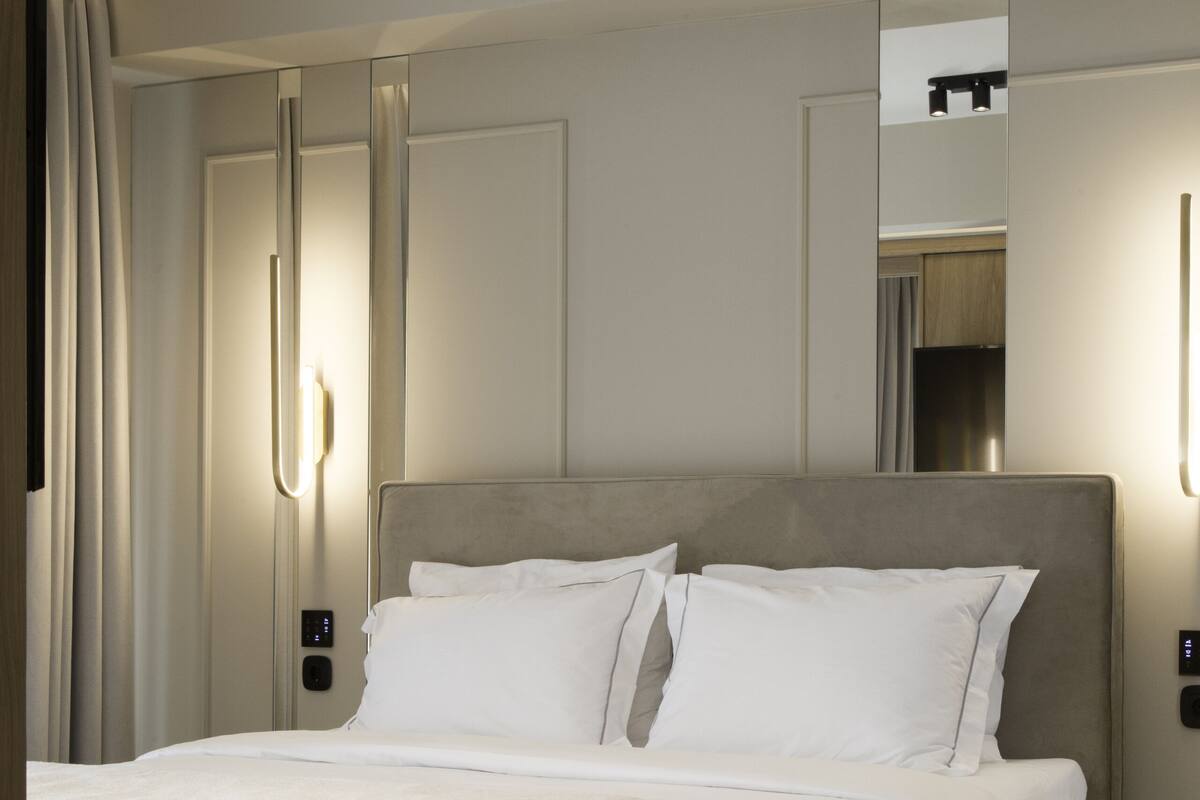 A modern hotel room with a large bed, a bedside table, a mirror, and a wall-mounted light fixture.