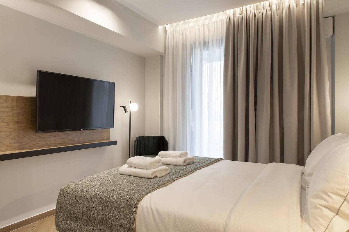 A modern hotel room with a flat-screen TV mounted on the wall, a bed with a grey textured bedspread, white pillows, and a set of neatly folded white towels on the bed.