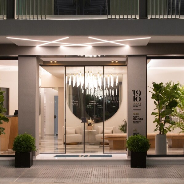 A modern hotel entrance with a glass facade, a chandelier, and a reception desk.
