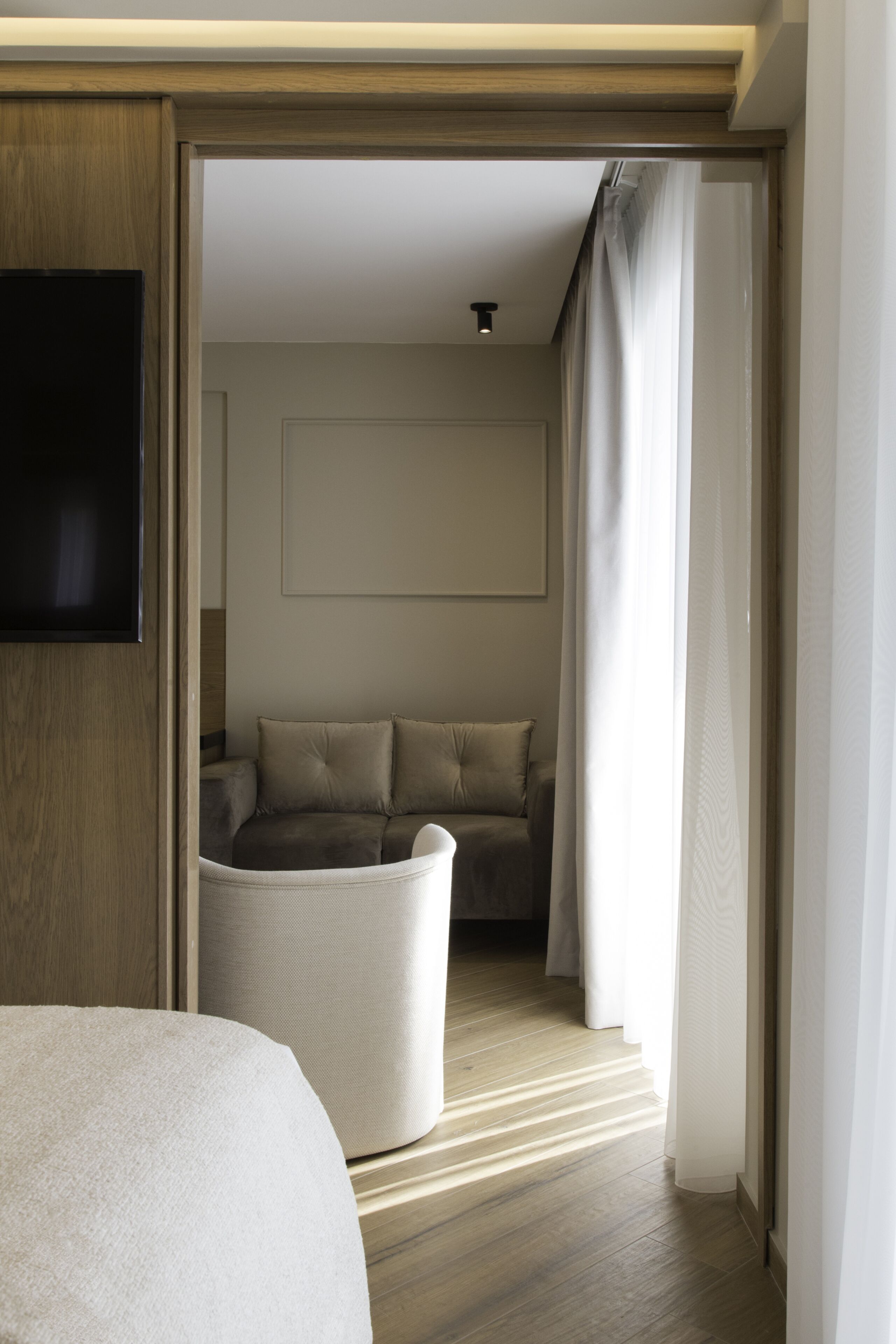 A modern hotel room with a wooden floor, a large bed, a seating area with a sofa and cushions, and a flat-screen TV mounted on the wall.