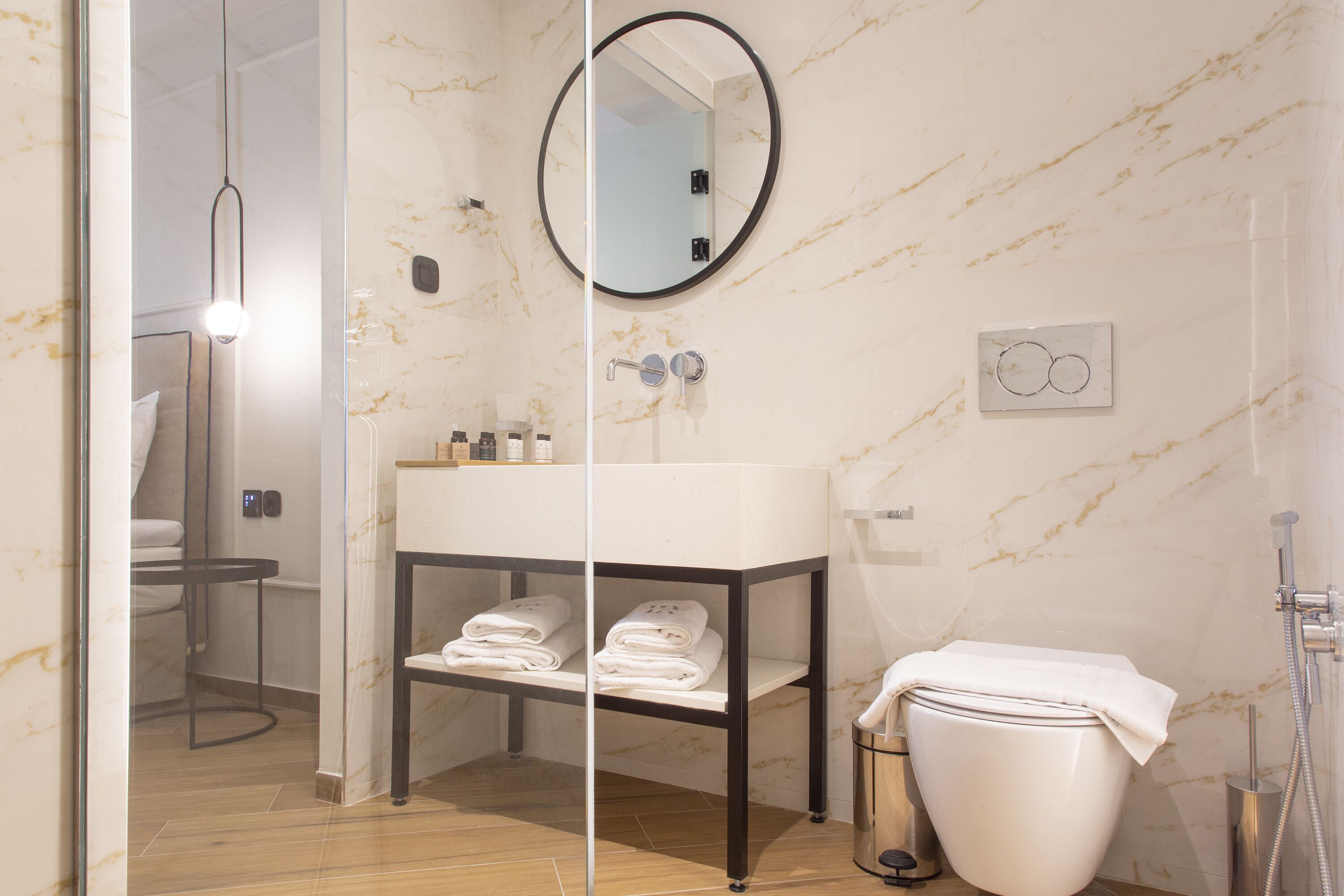 Un baño moderno con pared de mármol, un espejo redondo, un lavabo con toallero y un inodoro con ducha.