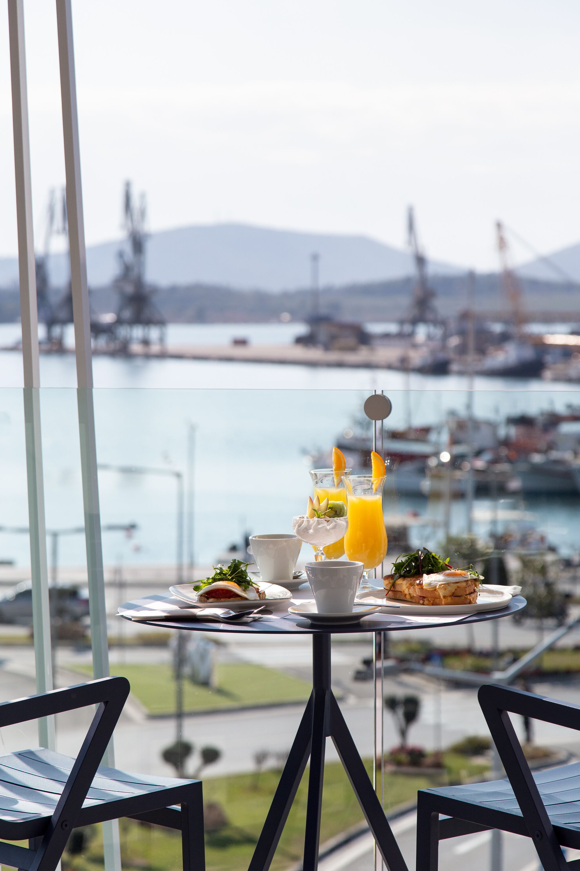 Una mesa con comida y bebidas, con vistas a un puerto deportivo con barcos y una montaña distante.