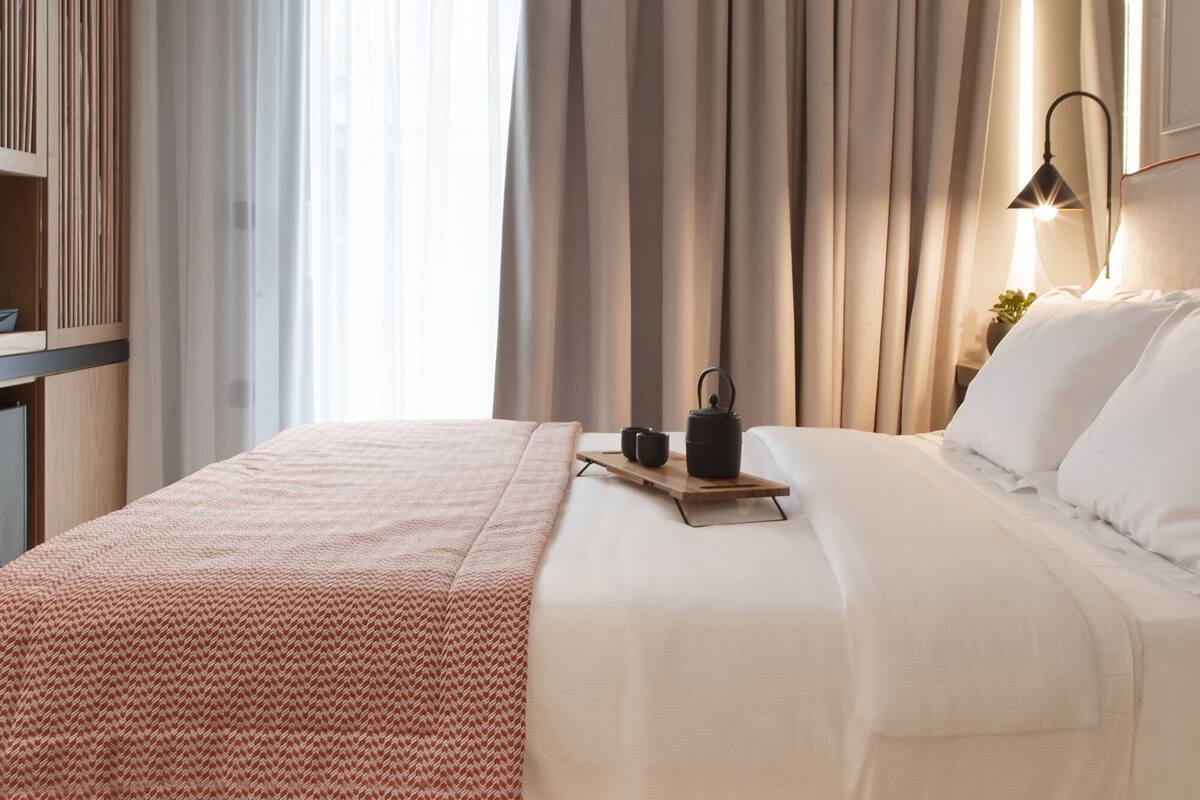 A neatly made bed with a red patterned blanket, a bedside table with a lamp, and a window with sheer curtains.