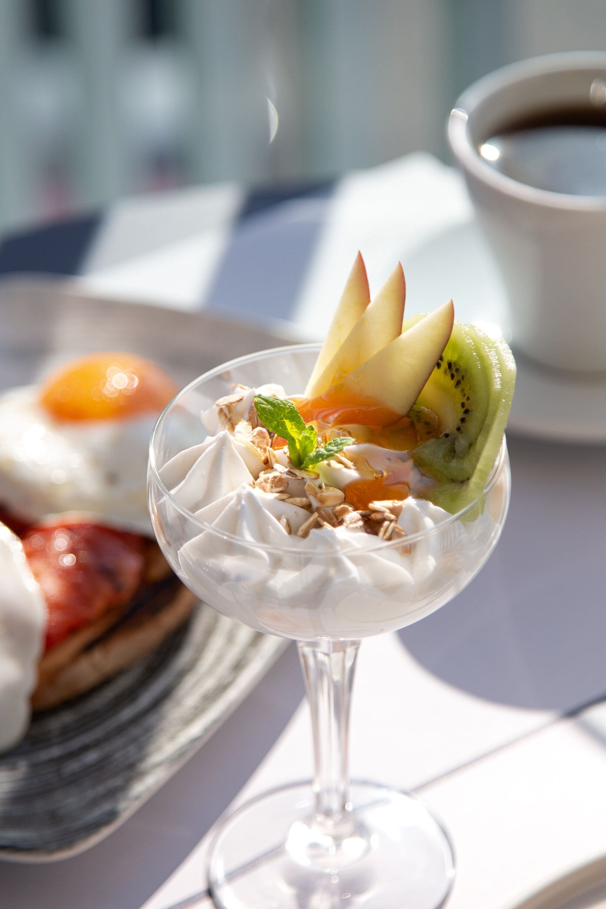 Dessert i et glas med flødeskum, frugt og granola, en tallerken med et wienerbrød, og en kop kaffe på et bord.