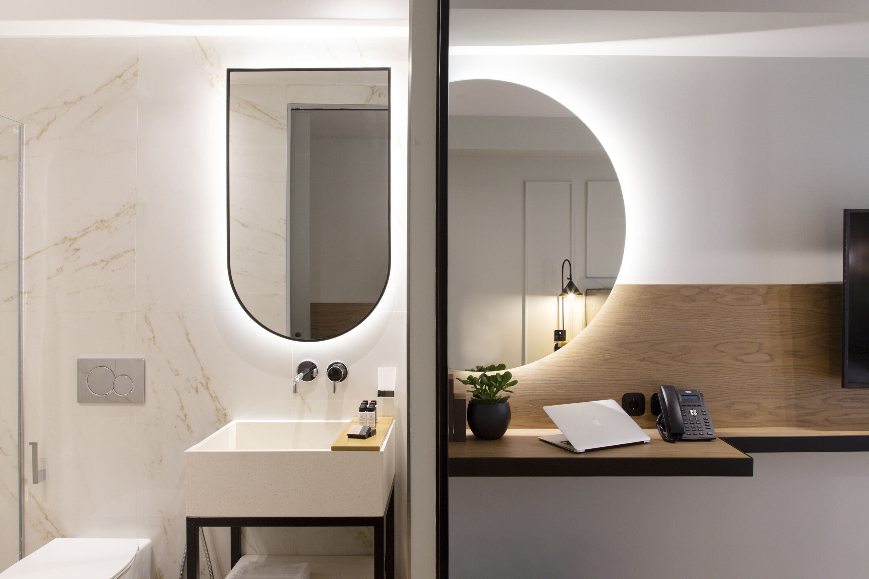 Un baño moderno con lavamanos de mármol, un espejo grande y un tocador de madera con un teléfono y una planta.