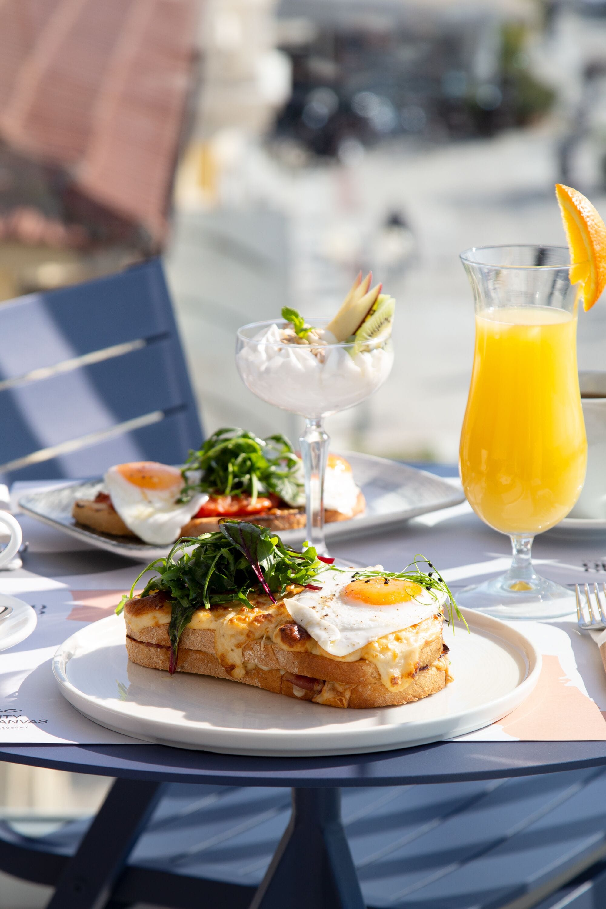 A table with a plate of food, a glass of orange juice, and a dessert on a sunny outdoor setting.