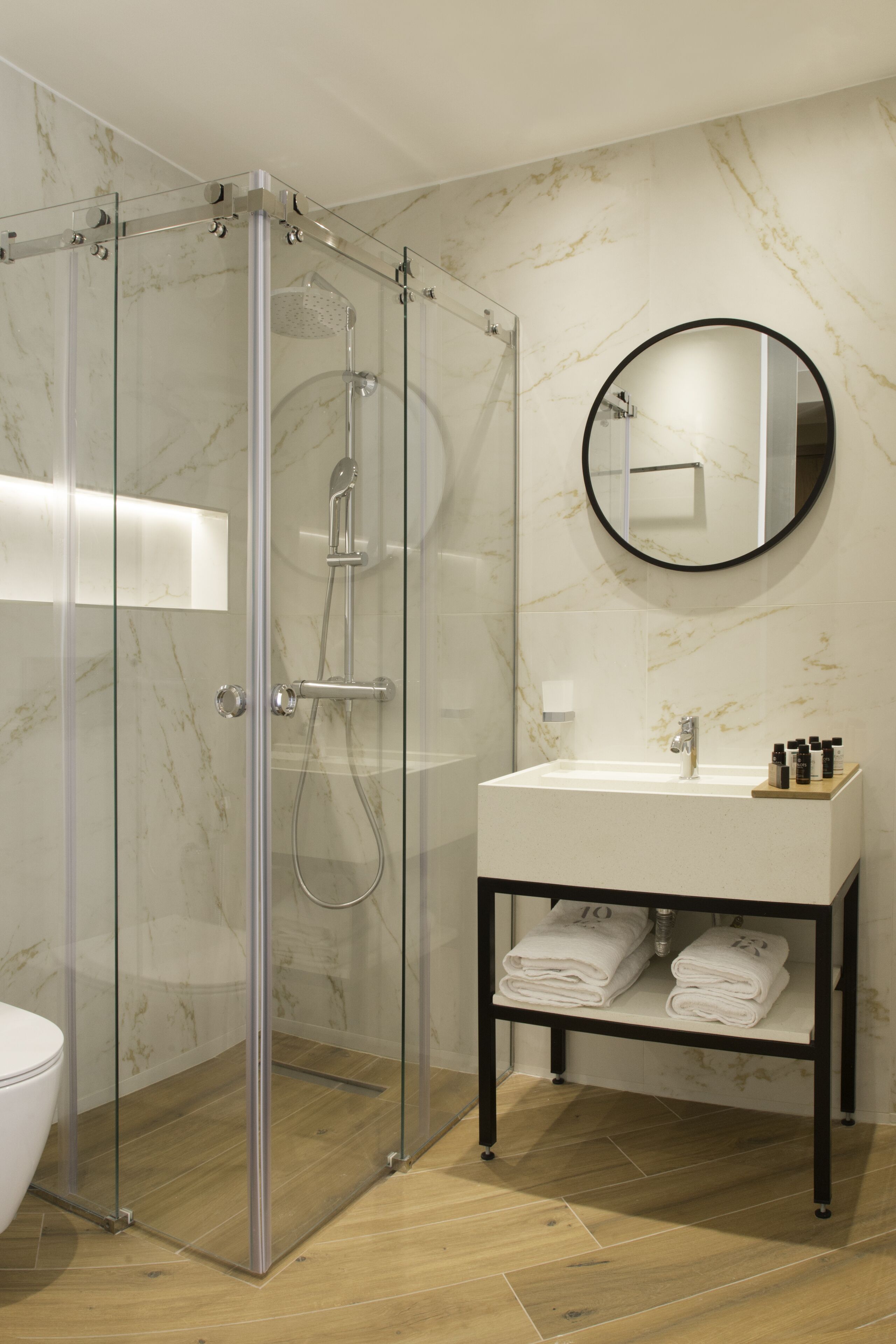 A modern bathroom with a glass-enclosed shower, a round mirror, a white sink, and neatly folded towels on a shelf.