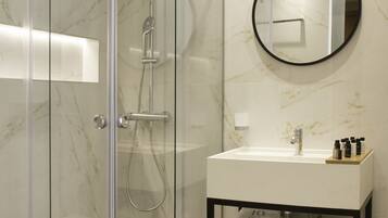 A modern bathroom with a glass-enclosed shower, a round mirror, a white sink, and neatly folded towels on a shelf.