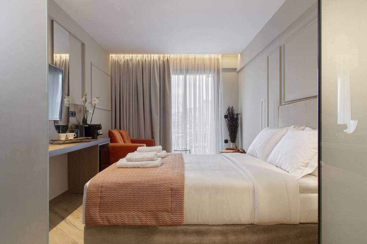 A modern hotel room with a large bed, a desk with a chair, a mirror, and a view of the city through the window.