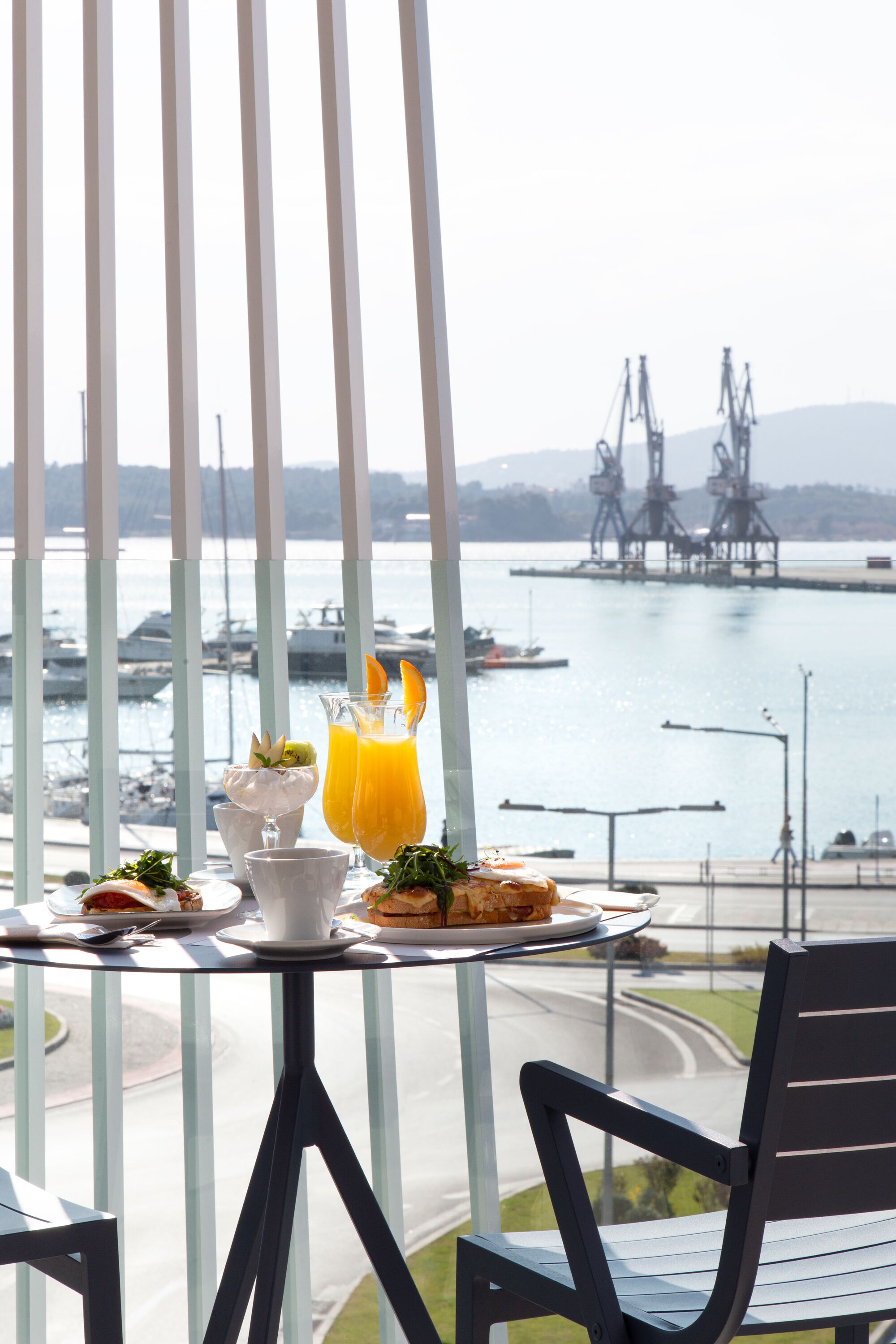 Un balcón con una mesa lista para el desayuno, con vista a una marina que incluye barcos y estructuras industriales.