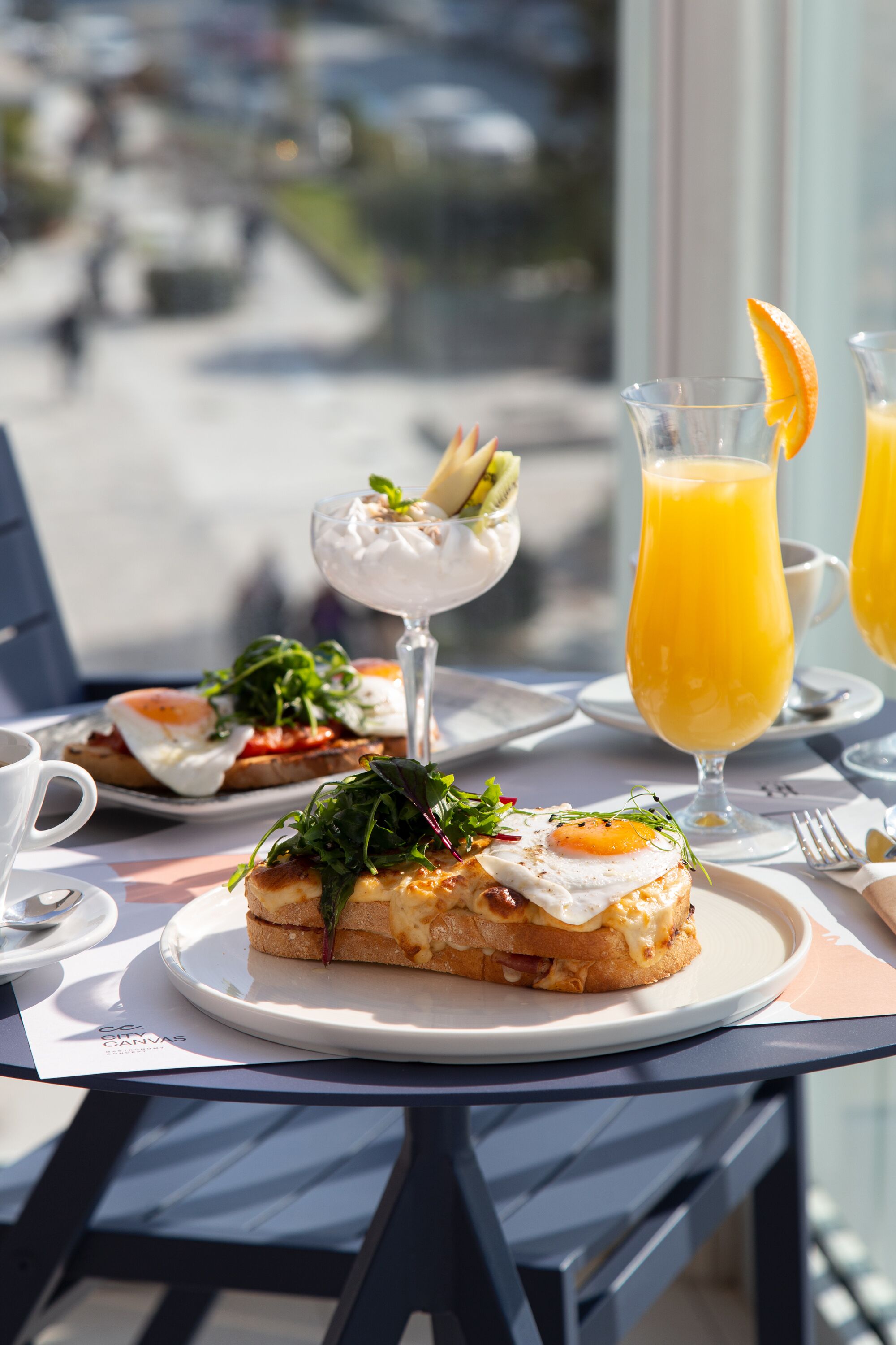 A table with breakfast items including toast with egg and greens, a glass of orange juice, and a dessert with a fruit garnish.